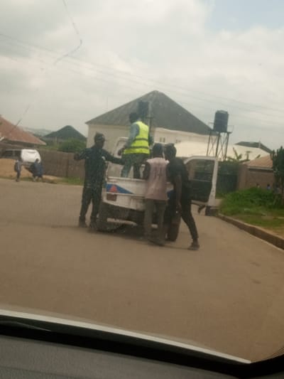 In Bar Jirgi Summa, Nigeria, a white pickup truck with its doors open is parked on a paved road. Several individuals are gathered around the truck, some appearing to be loading or unloading items. One person in a bright yellow vest is standing in the back of the truck. Another person in a dark shirt stands near the driver's side door. Two other individuals are near the rear of the truck, one wearing a light-colored shirt with writing on it and the other in a dark shirt. In the background, there are buildings with pitched roofs, a water tower on a metal frame, and vegetation. The sky is overcast. The scene suggests an activity involving transport and possibly manual labor.