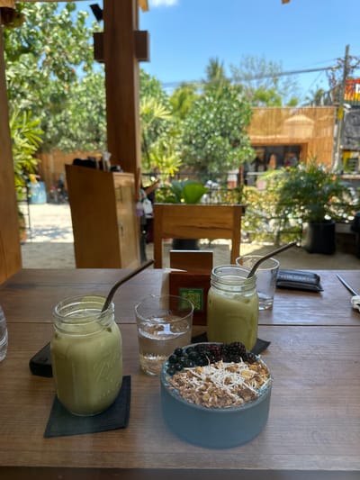 The image shows a wooden table in an outdoor setting, likely a cafe or restaurant. There are two mason jar glasses filled with a pale green liquid, possibly smoothies or juice, each with a stainless steel straw. Between them are two clear glasses, one containing water. To the right is a blue bowl filled with what appears to be a mix of granola, shredded coconut, and various berries. Each glass is placed on a square black coaster. The background includes greenery, suggesting a tropical environment, and parts of wooden structures. People can be seen in the distant background, but they are not clearly identifiable. The image is most likely taken in Puerto Escondido, Mexico.