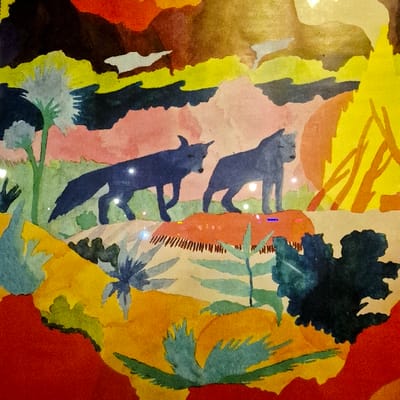 This image displays a stylized artwork, likely a watercolor or gouache painting, observed in the city of Strasbourg, France. The central subjects are two dark blue, canid-like animals, possibly wolves or foxes, depicted in profile and moving from left to right across a raised terrain. The lead animal on the left has prominent pointed ears and is captured mid-stride with a front leg lifted. The second animal, positioned slightly behind and to the right, appears to be standing or initiating movement.

The landscape is rendered with abstract, organic forms in a rich, multi-layered color palette. The animals stand on a light brown landmass with areas of reddish-orange texture. The immediate foreground features undulating shapes in red, orange, and dark green, along with stylized light blue-green spiky foliage. In the midground, a tall, light blue-green plant with a fluffy top is visible on the left, and green leafy plants emerge from the terrain. The background consists of successive layers of pale pink, dark blue, and yellow-brown forms, suggesting distant hills or atmospheric phenomena. A prominent, intensely bright yellow, vertically oriented shape on the far right resembles a stylized fire or a powerful light source. The artwork's surface displays visible reflections, indicating it is likely behind glass or a protective layer.