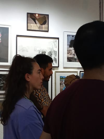 A group of people stand indoors, presumably at an art exhibition, as evidenced by the framed pictures on the wall. A woman in a blue shirt and a man in a patterned shirt are in the foreground, partly obscured by another person in a burgundy shirt. The wall features several framed artworks, including a high-heeled shoe, an abstract piece, and landscape photographs.
