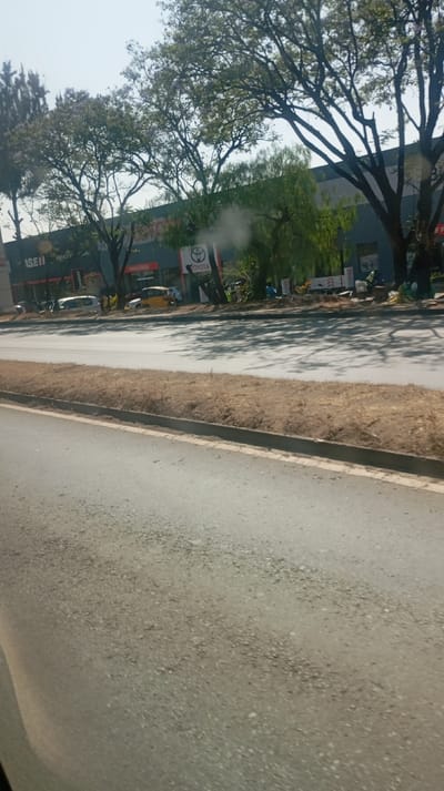 A street view captures a wide asphalt road with a raised median of dry grass and a curb. Across the road, trees line a building facade, partly obscuring signs reading "ISEII" and "TOYOTA." Vehicles, including a yellow tuk-tuk, are parked along the street. The sky is bright, and the scene is bathed in daylight.