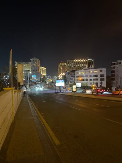 A night scene depicts a city street illuminated by streetlights and building lights. Several high-rise buildings line the street, with traffic moving in both directions. Billboards are visible alongside the road. The sidewalk is adjacent to the street, with a barrier separating them. The scene is captured from an elevated position, giving a perspective down the street.