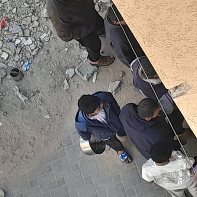 Aerial view of a group of people near a building. A person in a blue jacket holds a metal container. The ground is a mix of pavement and debris. The location is Khan Yunis, Palestinian Territory.