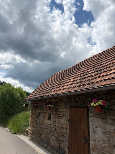 The image, taken in Wisła, Poland, showcases a rustic building under a dynamic, partly cloudy sky. The building features a steeply pitched roof covered in numerous reddish-brown tiles, laid in neat, overlapping rows. A dark grey gutter runs along the edge of the roof. The walls below are constructed from rough-hewn, irregular stones, lending a traditional, sturdy character to the structure.
Several terracotta-colored pots, brimming with vibrant hanging flowers in shades of pink, yellow, white, and purple, are attached to the stone wall, adding splashes of color. On the upper part of the stone facade, a dark wooden door or shutter is visible, secured with a metal latch. Below it, there's a small, rectangular opening, possibly a window, and another wooden element.
The sky dominates the upper and left portions of the photograph, presenting a mix of weather conditions. Areas of clear blue sky are interspersed with fluffy white clouds, especially in the upper left corner. Towards the middle and lower left, the clouds become denser and darker, creating a dramatic atmospheric effect.
In the lower-left and middle sections of the image, lush green vegetation, including trees and grass, can be seen. A grey, paved road or pathway curves along the very bottom right edge of the frame, bordering the grassy area. The scene is illuminated by bright daylight. There are no people visible, nor any discernible text. The image conveys a sense of traditional architecture nestled within a natural, slightly dramatic landscape.
