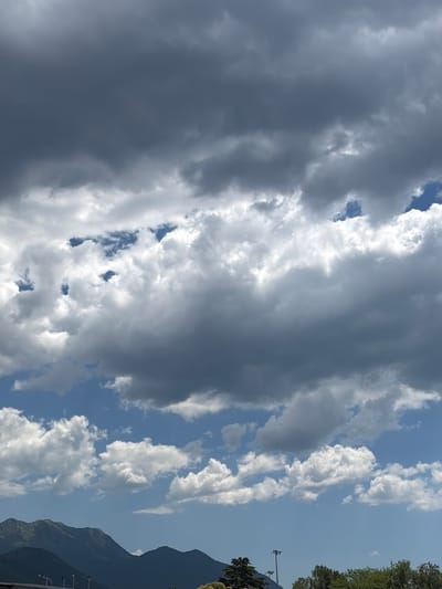 A wide shot captures a mountainous landscape under a dramatic sky. The lower third of the image shows dark, silhouetted mountain ranges. In the foreground, a few trees and streetlights are visible, suggesting a semi-urban or edge-of-town setting. The upper two-thirds of the image are dominated by a dynamic sky filled with a mix of dark, heavy clouds and brighter, wispy formations, with patches of blue sky visible in between. The overall impression is one of changing weather, possibly before or after a storm. The location is identified as Mrčevac, Montenegro. No people or specific actions are depicted.