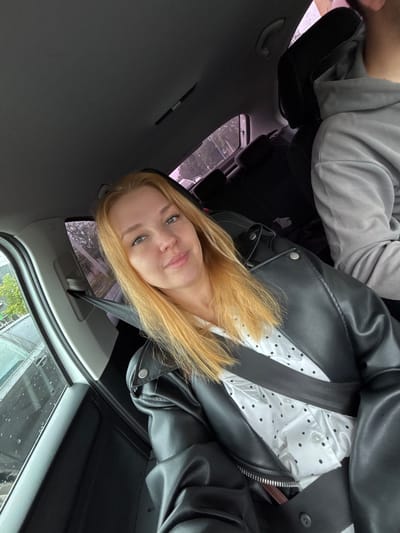 A blonde woman with fair skin and green eyes is captured in a selfie taken from inside a car. She is wearing a black leather jacket over a white shirt with black polka dots. A black seatbelt is fastened across her chest. The interior of the car is visible, including a dark-colored headrest and a grey hooded sweatshirt of another person in the passenger seat. Outside the car window, raindrops are visible on the glass, suggesting inclement weather. The background shows trees and possibly buildings, indicating an urban or suburban setting. The scene appears to be in the city of Zavolzhye, Russia.