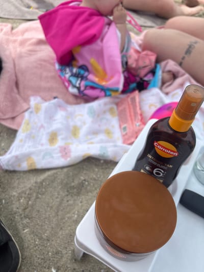 The image depicts a close-up view of beach essentials on the sand. In the foreground, a round, brown jar with a metallic sheen sits on a white plastic tray. To the right of the jar is a dark brown bottle with an orange spray nozzle, identified by the "Carroten INTENSE TAN" label. The bottle has text indicating "Tanning Oil Spray and Spray Ha Mavore". The tray and bottle are positioned on a sandy surface, with a piece of a black sandal visible on the far left. In the background, a pink towel and a pink baby blanket with cartoon characters are spread out, suggesting a family outing. There are blurred figures of people in the background, including legs and a baby's arm. The setting appears to be a beach, possibly in Varna, Bulgaria, given the context. The overall scene is casual and suggests a day spent at the seaside.