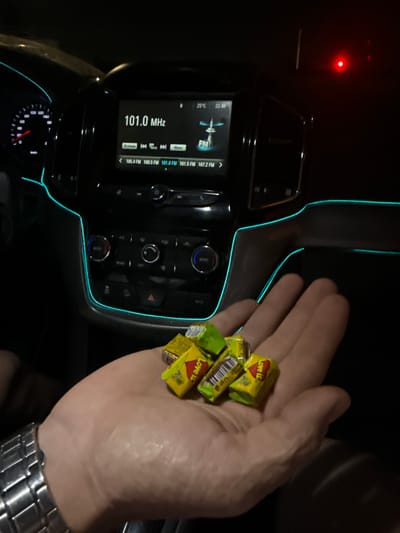 The image shows a person's hand holding a small pile of yellow candies, each wrapped in brightly colored packaging with the "Love is..." logo. The candies are mostly yellow with green and red accents on the wrappers, and the "Love is..." text is clearly visible in red. The hand appears to be an adult's, with a silver watch on the wrist.
The background is the interior of a car at night. The car's dashboard is visible, with a car stereo system displaying "101.0 MHz" and other radio station frequencies. There is also a speedometer with a needle pointing to a low speed. The car's interior is illuminated by subtle blue ambient lighting along the dashboard trim, and a small red indicator light is visible in the upper left corner. The overall scene suggests the person is in a car, likely driving or parked, at night. The focus is on the hand and the candies, with the car interior serving as context. No specific emotions are conveyed beyond the implied enjoyment of the candies. The location cue "Tashkent, Uzbekistan" is provided but not visually discernible in the image itself.