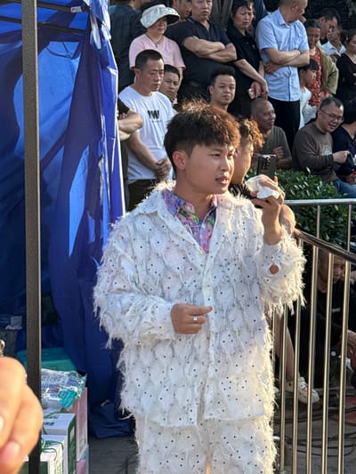 The image shows a crowded outdoor scene in the city of Shapingba District, China. A person in a white, feathery outfit is in the foreground, with a blue tarp beneath. A metal railing separates the individual from the crowd, and various boxed items are visible. Many individuals lie close together on the ground, looking upwards, potentially at the person in white. Another person holds up a cell phone to potentially record the subject in the white feathery outfit.