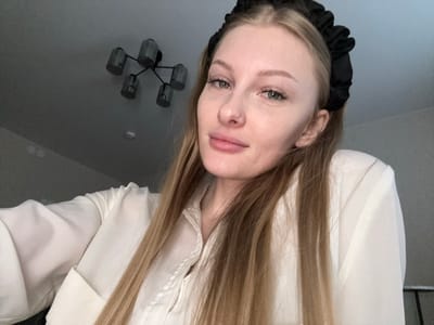 A young woman with blonde hair and a black scrunchie headband is pictured from the chest up, looking towards the camera. She is wearing a white collared shirt with buttons. The background is a plain wall with a dark ceiling fixture consisting of multiple cylindrical lights. The setting does not provide any specific visual indicators of its location within the city of Perm, Russia.