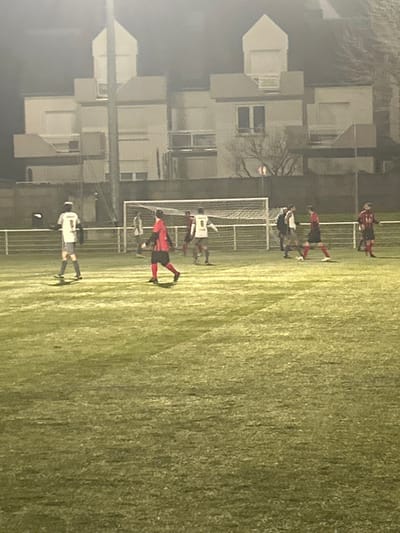 A soccer game unfolds on a green field. Players in red and white jerseys surround a net. Some players are actively engaged, while others stand on the sidelines. Behind the soccer field, a row of white buildings adds to the backdrop. The light suggests it might be evening or night.