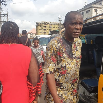 A daytime street scene in Onitsha, Nigeria, depicts an active urban environment with multiple pedestrians and vehicles. In the foreground, a bald adult male, appearing to be middle-aged, faces slightly right with a neutral to serious expression. He wears a short-sleeved traditional African patterned tunic in shades of brown, gold, red, and black, featuring floral and swirl motifs. To his left, the back of an individual in a bright red dress with dark braided hair is visible. Their right arm, covered by a black sheer sleeve with red horizontal trim, extends into the frame.
Further into the midground, an adult female with short, dark hair walks towards the viewer, wearing a patterned black and white top with a red floral detail pinned to the chest, and a black and red striped skirt. Behind her, the back of an adult male in a black shirt with a dark satchel slung across his body is seen.
The background features multiple white vans or minibuses parked or in motion. On the far right, part of a yellow commercial vehicle, possibly a Keke Napep or a taxi, is visible. Multi-story buildings, some appearing weathered and aged, line the street under an overcast sky. Overhead utility lines and power poles are present throughout the upper portion of the scene. The ground surface appears to be paved, typical of a busy city street.