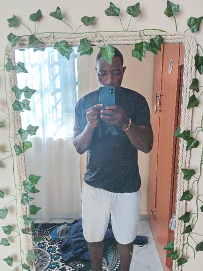 A person in a black shirt and white shorts takes a mirror selfie. The mirror has a rope border with artificial ivy leaves attached. Behind the person, reflected in the mirror, is a wooden cabinet above a window. The person's legs are visible, standing on a floor with a patterned rug and some blue clothing items. The scene is brightly lit by daylight filtering through the window.
