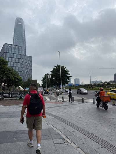 The image depicts a street scene in Haizhu District, China, under a cloudy sky. In the foreground, a man wearing a red t-shirt and khaki shorts walks away from the camera, carrying a black backpack and holding a bottle in his right hand and a mobile phone in his left. He is walking on a gray tiled sidewalk with tactile paving.
Further ahead, a pedestrian crossing is visible. Several people are crossing the street, including a woman with a backpack and another person in white. On the road, a scooter with an orange delivery box is being ridden by a person in an orange jacket. A yellow taxi is also visible.
The background features modern buildings, including a tall, curved skyscraper with a distinct top, and a multi-story glass-fronted building. Trees line the street, and there are streetlights and traffic signals. In the distance, more buildings and construction cranes suggest a developing urban area. The overall atmosphere is that of a typical city day with moderate activity.