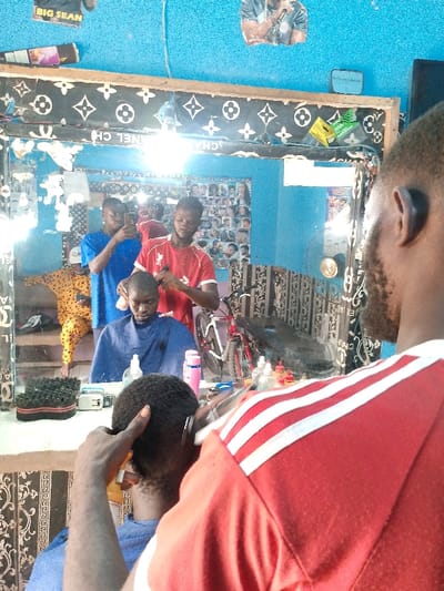 This image depicts the interior of a barbershop located in Neni, Nigeria. The vantage point is from behind a male client receiving a haircut, facing a large, rectangular mirror that reflects the main activity within the establishment.
In the immediate foreground, the back of a male individual's head is visible, showing short, dark, curly hair. An implement, consistent with a hair clipper, is positioned near the top of the head, operated by an unseen hand. The individual wears a red shirt with three white stripes across the right shoulder.
The mirror reflects the following: A young male barber, wearing a red t-shirt with 'LFC' text on it, is actively cutting the hair of a male client. This client is seated in a barber's chair, draped in a blue barber's cape, and looks forward. Standing to the left of the barber, another young male, dressed in a blue t-shirt, holds a smartphone pointed towards the mirror, seemingly capturing an image or video. Further in the reflected background, a fourth individual is seated, wearing a yellow and brown patterned garment.
The barbershop walls are painted a textured light blue. Above the mirror, several posters are affixed, one prominently featuring the text 'BIG SEAN'. The mirror itself is enclosed by a black frame adorned with repeating white patterns resembling the Louis Vuitton monogram. A single bare light bulb hangs centrally above the mirror, producing a significant glare reflection. A counter situated directly below the mirror holds various items, including multiple bottles of hair products (some with red caps), a pink plastic bottle, a clear dispenser, and a black hair sponge. The wall segment beneath this counter is covered with black wallpaper featuring a white Greek key pattern. In the reflected background, a chart displaying various haircut styles is visible on a wall, and a white bicycle with red tires is leaning against another wall.