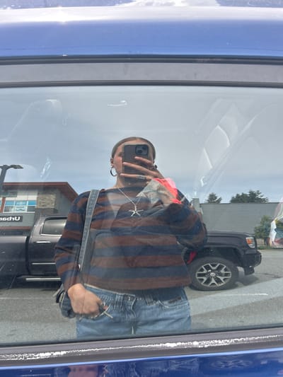 A reflection is captured in a vehicle window. The reflection depicts an individual holding a smartphone and wearing a striped shirt, blue jeans, and a vest. Also reflected are a vehicle with large tires, another car, a building with windows, and trees. The specific location cannot be determined based on the image alone.