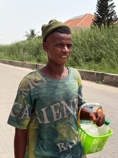A young man stands outdoors on a paved surface, possibly a road or sidewalk, in Lekki, Nigeria. He is holding a green plastic basket filled with what appears to be bottled water and other items. The man is wearing a green t-shirt with large white lettering and a matching green beanie. The background includes lush green vegetation, a building with a red roof and solar panels, and a concrete curb. The weather appears sunny and clear.