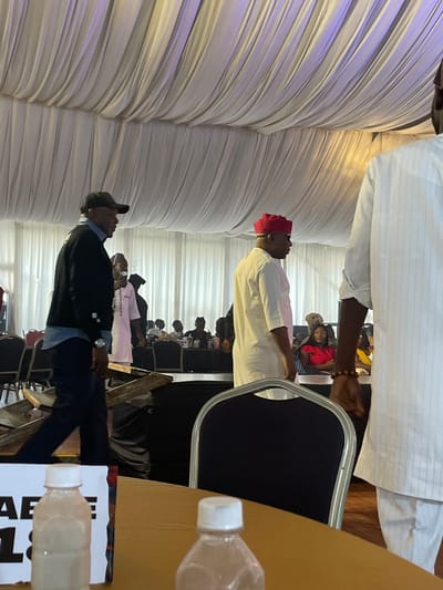 A medium shot captures an indoor event, likely a gathering or conference, within what appears to be a banquet hall in Barakin Lerawi, Nigeria. The ceiling is draped with white fabric, creating a textured canopy illuminated by soft, diffused light that casts purple and yellow hues. In the foreground, a table with a brown surface is partially visible, holding two plastic water bottles with white caps. A sign labeled "AE 18" is placed behind the left bottle.
The mid-ground features several individuals. A man on the left, wearing a black baseball cap, a blue collared shirt, and a black jacket, is walking towards the left edge of the frame. He has a watch on his left wrist. To his right, a man in a white traditional outfit and a red cap is standing and looking to his left. Behind them, other attendees are seated at tables, some partially obscured. A woman in a red top is visible in the background.
The background reveals more tables and chairs, with some individuals standing or walking. The overall atmosphere suggests a formal or semi-formal event. The lighting and drapes indicate a decorated interior space, possibly a hotel ballroom or event center. The presence of a sign with "AE 18" suggests a possible seating arrangement or event identifier.