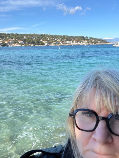 A selfie-style photograph depicts a person in the foreground against the coastal landscape of Antibes, France. The upper face of an individual with blonde and grey hair, wearing black-framed glasses, occupies the lower-right quadrant. Their right eye is visible and directed towards the camera. The person wears a dark, possibly quilted, jacket. The expansive midground is dominated by clear, multi-toned blue-green seawater, revealing subsurface details and exhibiting minor surface agitation. White mooring buoys are sparsely distributed across the water. The background presents the Cap d'Antibes peninsula, characterized by dense green foliage, numerous white residential structures, and a prominent white lighthouse accompanied by a slender tower on the highest elevation. Various boats are observed on the water near the distant shoreline, including a large white vessel on the far right. The horizon extends to reveal distant snow-capped mountains under a bright blue sky with minimal cloud cover. The scene is illuminated by natural sunlight.