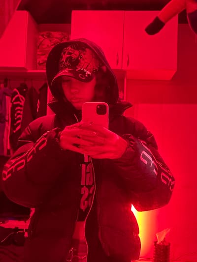 The image is a vertical mirror selfie, rotated 90 degrees clockwise from its natural orientation, taken in an indoor setting bathed predominantly in intense red light, with a strong yellow-orange glow emanating from the right background. While the location context is Buenos Aires, Argentina, no specific geographical cues are visible in the image itself.
The subject is a person whose face is largely obscured by a light-colored smartphone they are holding to take the picture. They are wearing a dark, possibly black, puffy jacket or hoodie. Visible on the jacket are large, light-colored letters such as "JG" or "JC" on the upper chest area, and similar lettering further down. The hoodie also features an intricate, dark design, possibly a skull or abstract pattern, on its front. Around the person's waist or hip area, a dark band with visible text, including the number "2" and other indistinct letters, is apparent, suggesting it might be part of their clothing or an elastic band. The person's visible hand, holding the smartphone with a dual-camera system, shows what appears to be detailed nail art or rings.
In the background, to the left, a light-colored, possibly white, appliance resembling a small refrigerator or a two-door cabinet is prominent, with a smaller cabinet or shelf unit below it. On the far left, a dark, plush object, possibly part of a stuffed animal, can be seen. On the far right, near the intense yellow-orange light, there is a textured, possibly woven object.
The primary activity depicted is the taking of a mirror selfie. The time of day is not discernible due to the artificial lighting. The pervasive red light creates a moody and high-contrast atmosphere, strikingly contrasting with the warm, bright yellow-orange light from the background. No specific emotions are discernible from the partially hidden face.