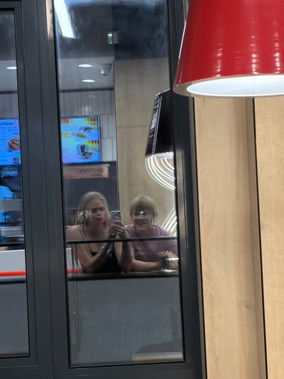 Two young women are reflected in a glass window. The woman on the left has blonde hair and is wearing a black tank top. She is holding a smartphone and appears to be taking a selfie. The woman on the right has short blonde hair and is wearing a pink shirt. She is looking at the woman on the left and smiling. They are seated at a counter, possibly in a fast-food restaurant, given the menu boards visible in the background. The restaurant appears to be located in Pervomaysky, Russia. The overall scene is casual and appears to be a moment of shared activity between friends.