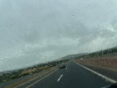 The image depicts a driver's perspective through a rain-streaked windshield, looking out at a wet asphalt road stretching into the distance. The sky is overcast and gray, with visible streaks of rain on the glass. The road has yellow and white lines, indicating a highway or major road. The surrounding environment appears to be a rural or semi-rural landscape with low hills covered in sparse vegetation. There is a dark-colored vehicle ahead on the road. The scene suggests a journey on a rainy day.