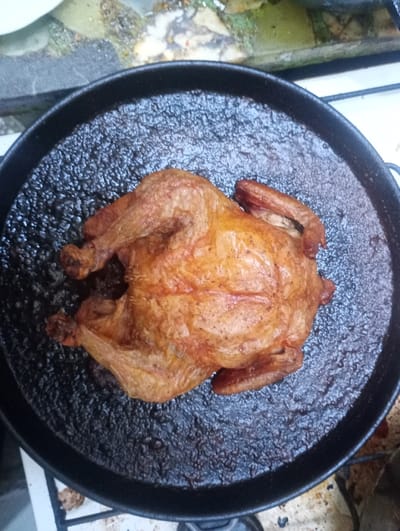 A whole roasted fowl, likely a chicken, is centrally positioned within a large, circular, black frying pan in Wilde, Argentina. The bird displays a golden-brown, crispy skin with some darker, caramelized areas, indicating it is fully cooked. The pan's interior surface is dark and textured, consistent with cooking residue or heavy seasoning. The pan rests upon a metal grate, part of a white-enameled stove, which shows signs of use. The background features a mottled, rough surface, possibly concrete or stone, with minor green organic growth and varied discoloration, suggesting an outdoor or semi-outdoor setting. No individuals or active interactions are visible in the scene.