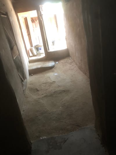The image depicts an indoor corridor with a dirt floor and rough, unfinished walls. In the background, there are two doorways leading to brighter areas, possibly outdoors. Through one doorway, a plastic chair and a bucket are visible. The overall impression is one of simplicity and possibly poverty. The location is identified as a residential area in Jalingo, Nigeria.