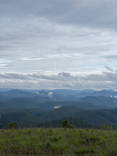 The image shows a mountainous landscape with a foreground of green vegetation and distant, hazy mountain ranges under a cloudy sky. The location is likely near Trường Thọ, Vietnam, based on the surrounding environment.