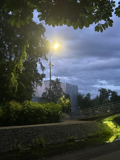 The image presents an outdoor scene captured at dusk or night in Kazan, Russia. The sky is heavily overcast with dark grey and blue clouds. Overhanging the upper portion of the frame are dark green leaves from trees, notably large palmate leaves consistent with horse chestnut. A single streetlamp, emitting a bright yellow light, stands prominently in the mid-ground, positioned to the left of the central axis. A white surveillance camera is mounted beneath the light fixture on the streetlamp pole.

Beneath the trees and extending to the left and center are dense, dark green bushes and trees. Behind this vegetation, a large, geometrically designed building with a light grey or white facade is visible; this structure is identified as the **Ekiyat Tatar State Puppet Theatre** in Kazan, Russia.

A narrow body of water, likely a canal or stream, traverses the lower part of the image. The water's surface reflects an intense, localized greenish-yellow light emanating from the right side of the frame, possibly from illumination beneath a pedestrian bridge. A metal pedestrian bridge with railings spans this water body on the right. A dark, rocky or gravel-covered bank separates the water from a paved pathway running parallel to the water. No individuals are present in the scene.