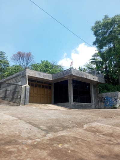The image shows a one-story building in Nawng Hkar, Myanmar, with a gray concrete structure, a tan garage door, and tinted windows on the right side. A gray fence is visible on the left. Graffiti adorns a wall on the right side of the building, next to some plants. An Indonesian flag is on the roof. The foreground shows a cracked concrete driveway. Trees and foliage surround the building. The sky is blue with scattered white clouds. Overhead power lines are visible.