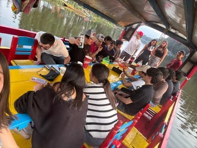The scene displays approximately fifteen individuals, predominantly young adults, on a traditional trajinera boat in the Xochimilco canals of Ciudad de México, Mexico. The boat's interior is brightly painted in red, yellow, and blue, featuring a central yellow table surface and perimeter benches. An overhead canopy covers the boat. On the table, multiple smartphones, a black crossbody bag, several red Coca-Cola cups, a green plastic bottle, and other beverage containers are present. One male in a grey baseball cap holds a clear liquor bottle aloft. Individuals are seen conversing, consuming beverages and snacks, and operating smartphones. The surrounding canal water is murky green, reflecting the sky. The background shows distant green vegetation, trees, and hills along the canal banks, with another boat partially visible.