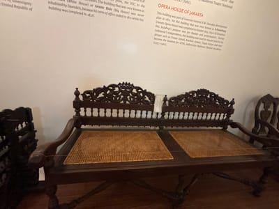 The image shows a bench with carved wooden armrests and a woven cane seat, set against a plain white wall. The bench appears to be part of a historical exhibit, as evidenced by the informational text on the wall to the right. The text, which is partially visible, discusses the "Opera House of Jakarta" and its history, including its development by Governor General H.W. Daendels and its completion in October 1814. The building's purpose for theater and performances, and its later use by Indonesian independence groups and theater practitioners, are also mentioned. The text indicates that the building became the location for ATNI, Indonesian National Theater Academy from 1957-1961. The overall scene suggests a museum or heritage site display in West Jakarta, Indonesia, focusing on historical architecture and cultural significance.