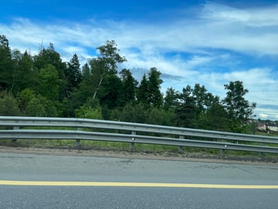 The image shows a view from a moving vehicle on a road in Бавыкино, Russia. The road is marked with a yellow line, and a metal guardrail separates the road from a forest. The sky is blue with scattered clouds. Some buildings can be seen in the distance.