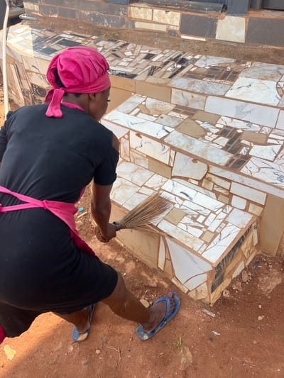A person is sweeping a mosaic tiled outdoor staircase. The person is wearing a black t-shirt, a pink headscarf, a pink apron, and blue flip-flops. The staircase is constructed from various sizes and shapes of mosaic tiles, including white, beige, black, and marbled tiles. The ground is composed of reddish-brown soil with scattered small rocks and dry vegetation. The location is identified as Nkpor, Nigeria, suggesting an outdoor setting in a community or residential area. The lighting suggests it is daytime.