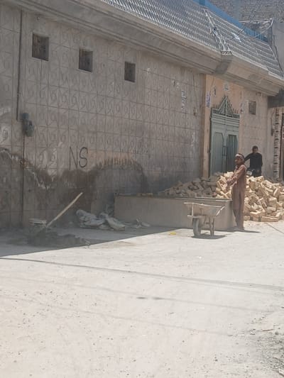 The image shows a street scene in Quetta City Tehsil, Pakistan. A building with a patterned facade occupies the left side of the frame. Three small square windows are visible on the lower portion of the wall. Above the windows, “NS” is written on the wall. Two men are near a pile of bricks: one man is wearing black clothing, the other is wearing tan clothing. A wheelbarrow is present. A long-handled tool rests against a wall. The street is clear, sunny, and devoid of traffic.