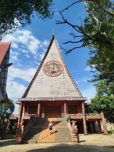 The image shows a unique building in An Phu Ward, Vietnam. A person is walking up the stairs to the entrance. The building has a triangular facade with a circular emblem in the center. The sky is blue with scattered clouds. Trees frame the building on both sides. The architectural style is distinctly local. A structure with a red roof is visible to the left.