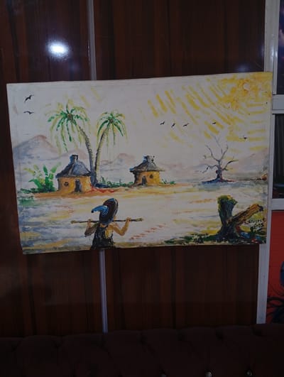 The image shows a painting depicting a rural scene in Adankolo, Nigeria. The painting includes:
* Two round huts with conical roofs.
* Two palm trees.
* A bare tree with several birds flying overhead.
* A person in the foreground carrying something (possibly a yoke) on their shoulders.
* An object that appears to be a tree stump in the foreground to the right.
* The painting is dated "2015".
* The background shows hills.
* Above the hills is a hazy or sunny sky with indications of a sun.
The painting is mounted on a wall with vertical wooden paneling. A brown leather sofa is in the foreground.