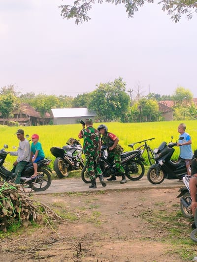 The image shows a scene in Karangmalang, Indonesia (latitude: -7.47, longitude: 111.064). There are four men standing near motorcycles. Two of the men are dressed in camouflage military attire. One man is holding a gun. The other man is wearing a red and black shirt. Two other men are seated on motorcycles. One man is wearing a blue shirt and the other is wearing a white shirt. There is a young boy wearing a red hat seated on the motorcycle being ridden by the man in the white shirt. All three motorcycles are black. There is also a green and black bicycle parked in the background. The scene is set in a rural area with green rice fields, trees, and a few buildings in the background. The ground is dirt and gravel. The overall atmosphere of the image is calm.