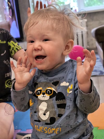 A close-up, eye-level, medium shot captures a toddler in a grey long-sleeved shirt with a raccoon graphic, looking to their left with an open mouth and raised hands, suggesting surprise or excitement. The toddler has blonde hair and is positioned in the foreground. Bubbles are visible in the air around the toddler. In the background, a portion of another child's arm and shoulder wearing a dark shirt with a lime green graphic is visible to the left. A pink sphere is partially visible behind the toddler to the right. The environment appears to be indoors, possibly a living room, with blurred background elements including furniture and windows. The lighting is natural and appears to be daylight. The image quality is somewhat soft with some grain. The location is not determinable from the image.