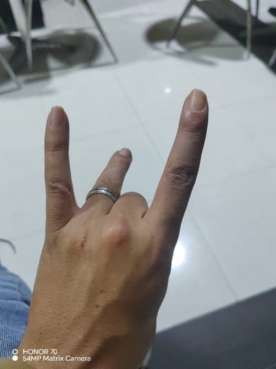 The image displays a close-up of a person's right hand performing the "rock and roll" hand gesture (corna gesture), with the index and pinky fingers extended upwards, and the thumb holding down the middle and ring fingers. A simple, silver-toned ring with two narrow bands is worn on the ring finger. The skin tone of the hand is medium, and some veins are subtly visible on the back of the hand. There is a small, reddish mark or blemish on the back of the hand, located between the knuckles of the index and middle fingers. The fingernails on the extended fingers are visible and appear natural, unpolished, and slightly grown out past the fingertips.

The background is out of focus, suggesting a shallow depth of field, with the hand being the primary subject. It appears to be an indoor setting with a light-colored, possibly tiled, floor. Reflections of metallic objects, possibly chair legs or furniture frames, are discernible on the floor. A distinct bright reflection from a light source is also visible on the floor in the mid-background. The overall lighting seems somewhat dim, but the hand is adequately lit.

In the bottom left corner of the image, a watermark is visible, which reads: "HONOR 70" and "54MP Matrix Camera."