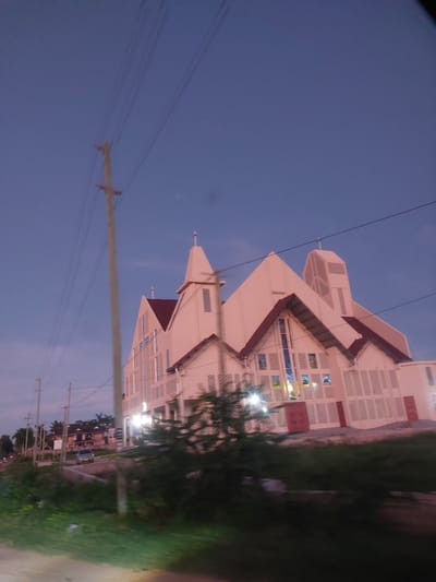 The image shows a tall, white church building with a series of tiered, angular roofs. The church has many windows, some of which are stained glass. In the foreground, there is a utility pole and wires running across the sky. To the right of the church is greenery, likely trees and vegetation. The sky is a dusky blue, suggesting it is either early morning or evening. A road is visible to the right edge of the image, next to the greenery.
The photo appears to be taken from a low angle, possibly from a moving vehicle. Based on the unique architectural style of the church, this is likely the Roman Catholic Holy Spirit Cathedral, located in Dar es-Salaam, Tanzania.