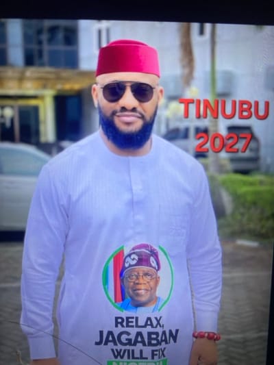 A man with a blue beard and sunglasses wears a red fez and a white shirt. The shirt features a button with a picture of a man wearing a purple cap, and the text "RELAX, JAGABAN WILL FIX NIGERIA". The text "TINUBU 2027" is visible in red above the man's head. He is wearing a beaded bracelet. The background is blurry, suggesting an outdoor setting.