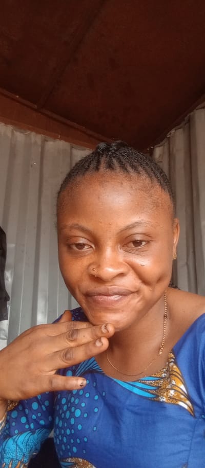 A close-up portrait shows a young woman with dark skin looking directly at the camera. She has her hair styled in small, neat braids pulled back from her forehead. A small, shiny nose stud is visible on her left nostril. Her expression is a soft smile, revealing dimples on her cheeks, and her eyes have a gentle, slightly downcast gaze. She wears a blue garment with a distinctive pattern featuring light blue circles and some yellow and brown designs on the sleeve and chest area. A thin, gold-colored chain necklace is visible around her neck, and a small, intricate gold-colored earring is in her left ear. Her right hand is resting with her fingers lightly touching her chin and lower jaw, and her fingernails appear unpolished.
The background behind her consists of a light grey or white corrugated metal wall or paneling, suggesting an enclosed or semi-enclosed structure. Above this, a dark brown or reddish wooden beam or ceiling structure is visible. The lighting is bright and natural, illuminating her face, suggesting it is daytime. No text is visible in the image.