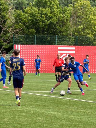 On a sunny day, a soccer game is underway on a green artificial turf field, situated in Esch-sur-Alzette, Luxembourg. Several players, all male, are actively engaged in the game. One player, wearing a dark blue jersey with the number 25 and the name "ASPORT," faces away from the camera. In front of him, other players in blue jerseys challenge for the ball. A referee in an orange shirt oversees the match. A red and white barrier stands in the background, and lush green trees border the field's edge.