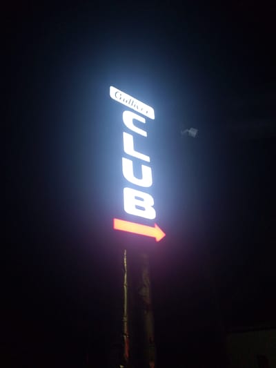 A vertical illuminated sign reads "Gulliver CLUB" in white LED letters, with "Gulliver" in a curved font at the top and "CLUB" in a sans-serif font below. An orange illuminated arrow points to the right from the bottom of the sign. The sign is attached to a pole that is partially visible. The background is dark, indicating it is nighttime. A small white security camera is visible mounted on the pole above the sign. The scene appears to be located in Bukhara, Uzbekistan, though no specific landmarks are visible.