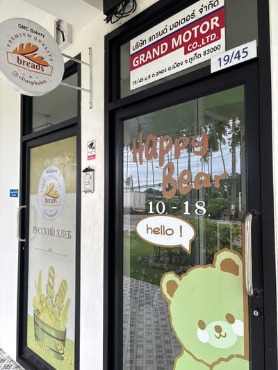 The image shows the exterior of a business in Chalong, Thailand. On the left, there is a circular sign for "GMC Bakery PREMUM QUALITY breads". Below this sign, the text "РУССКИЙ ХЛЕБ" (Russian Bread) is visible on the glass door, along with a graphic of bread loaves and wheat stalks in a wooden bucket. A small blue sign indicating "Staff Only" is also present on this door.
To the right of the bakery door, there is another entrance with a black frame. Above this door, a sign reads "GRAND MOTOR CO., LTD." in large red letters, with Thai text above and below. The address "19/45 ม.8 ต.ฉลอง อ.เมือง จ.ภูเก็ต 83000" (19/45 M.8, Chalong Subdistrict, Mueang Phuket District, Phuket 83000) is also visible on a white placard.
On the glass of the right-hand door, there is decorative text that appears to say "Happy Bear 10 - 18" in orange lettering, with a white speech bubble containing "hello!". A large graphic of a light green cartoon bear is also visible on this door, partially obscuring the interior view.
Additional details visible include a security camera sticker, a PEA (Provincial Electricity Authority) information box, and reflections of palm trees and a cloudy sky in the glass. The overall environment suggests a commercial street scene.