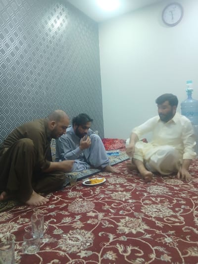 The image shows three adult males sitting on a carpet indoors. One man is wearing a light-colored traditional shalwar kameez and is kneeling. Another man is wearing a grey shalwar kameez and is sitting cross-legged, apparently eating something. The third man is wearing a brown shalwar kameez and is also sitting cross-legged. A plate with food is visible near the second man. Several empty drinking glasses are on the carpet nearby. A water dispenser and a clock are visible on the wall in the background. The wall has wallpaper with a wavy pattern.

Based on the clothing and cultural context, this scene is likely taking place in Quetta City Tehsil, Pakistan.