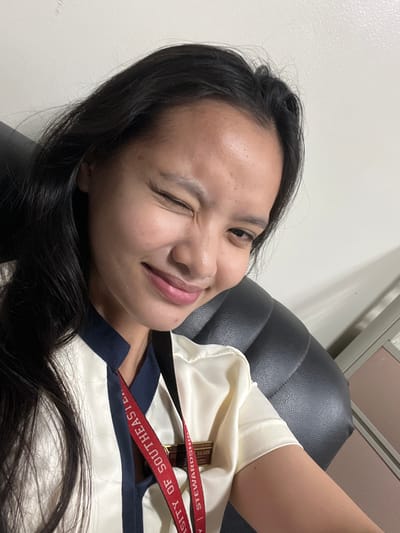A young woman is pictured in a selfie taken indoors, likely in Davao City, Philippines, as indicated by the lanyard around her neck which reads "UNIVERSITY OF SOUTHEASTERN PHILIPPINES." She is seated in a black leather chair, her head tilted slightly to her right, with her right eye closed in a wink and her left eye open. Her dark hair is long and falls over her left shoulder. She is wearing a uniform consisting of a cream-colored shirt with a navy blue collar and trim. The red lanyard is visible around her neck, featuring white text. The background appears to be a plain white wall with some subtle shadows, suggesting an indoor setting with artificial lighting. There are no other people or significant objects in the immediate vicinity. The overall impression is a casual, candid photograph.