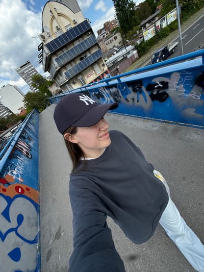 A person wearing a dark gray sweatshirt, white pants, and a dark blue baseball cap with a white "NY" logo stands on an overpass in Vozdovac Urban Municipality, Serbia. The overpass has blue metal railings covered in graffiti. Behind the person are buildings, including one with multiple solar panels, and a street with cars. The sky is cloudy with patches of blue.